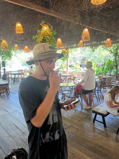 The image depicts an outdoor restaurant or cafe with a thatched roof covered in hanging vines and decorated with numerous woven bamboo pendant lights. Wooden floorboards run throughout the space, with tables and chairs scattered around.
In the foreground, a young person wearing a dark t-shirt and a light-colored bucket hat is captured in profile, holding a smartphone in their left hand and bringing a piece of food, possibly a slice of cucumber, to their mouth with their right hand. They are looking to their right.
In the midground, a woman with blonde hair, wearing a loose white top and dark shorts, stands facing away from the camera, resting her left hand on the back of a chair. Behind her, other diners are visible at tables. To the right, a young girl in a striped dress is seated at a wooden bench, looking down at something on the table.
The background reveals lush green foliage and trees, suggesting a tropical or semi-tropical setting. The overall atmosphere appears relaxed and casual, typical of a cafe or restaurant in a warm climate. While the exact location is not specified in the image, the presence of tropical vegetation and the architectural style of the lighting and roofing could be consistent with a location in Vĩnh Hải, Vietnam, known for its coastal and natural attractions.
