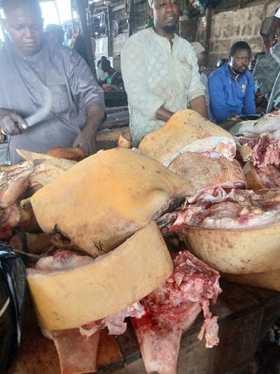 A butcher block displays raw meat, including sections of pig with skin, fat, and bloody cuts. Three men are visible near the meat, one with a cleaver, another in a white robe, and a third in a blue shirt. The scene appears to be in an open-air market.
