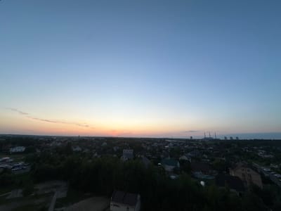 This aerial, wide-angle photograph captures a twilight scene over the Shchomyslitski Sel'ski Savet in Belarus. The sky displays a gradient of colors, transitioning from a pale blue overhead to soft oranges and pinks near the horizon. In the distance, a complex of industrial cooling towers and power plant structures is visible against the fading light. The foreground and midground are dominated by a residential area characterized by numerous houses with pitched roofs, interspersed with dense green trees. Roads and parked vehicles, including cars and vans, are present in several locations throughout the settlement. The overall impression is of a suburban or semi-rural landscape at dusk.