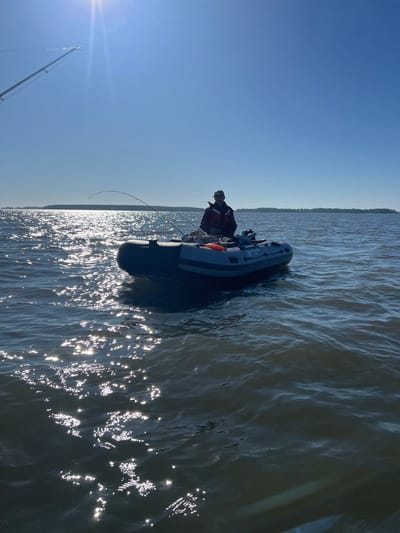A person wearing a life vest and a cap is seated in a gray inflatable boat on a body of water. The person is holding a fishing rod that is bent, indicating a possible catch. The boat is equipped with an outboard motor at the stern. The water is choppy with sunlight glinting off the surface. In the background, a distant shoreline with trees is visible under a clear blue sky. The scene appears to be a fishing excursion. There is no discernible information within the image that places the location in Novopichugovo, Russia.