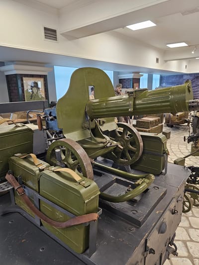 The image shows a mounted Maxim gun in a museum, possibly the museum located in Архангельское, Russia. The gun is painted olive green, and is mounted on a wheeled carriage. Ammunition boxes are attached to the carriage with leather straps. The background shows a hallway with artwork and a stone wall.