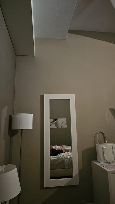 The image depicts a dimly lit room, likely a bedroom, with a neutral color palette dominated by beige walls and white accents. In the foreground, to the left, a floor lamp with a white cylindrical shade stands tall. To its right, a full-length mirror with a white frame is positioned. Reflected in the mirror, a person is lying on a bed, dressed in dark shorts and a maroon shirt. The bed is covered in white sheets. Above the bed, a rectangular artwork featuring a floral motif is visible on the wall. To the far right, a white tote bag with "LIU-JO" branding rests on top of a white surface, possibly a dresser or nightstand. The ceiling features architectural elements, appearing to be a raised section with a subtle texture. The overall impression is one of a private, potentially domestic space, suggesting the scene is set within a hotel room or a residential apartment in Alhama de Aragón, Spain, given the context provided.