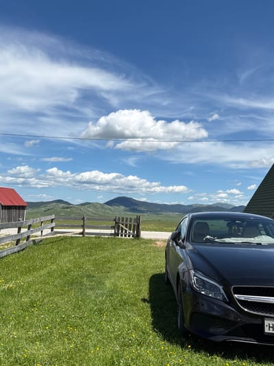 The image depicts a rural landscape under a bright blue sky with scattered white clouds. A black car is parked on a grassy area in the foreground. To the left, a rustic wooden fence with a gate encloses a section of the grass, and a small wooden building with a red roof is visible behind the fence. In the background, rolling green hills and mountains stretch towards the horizon. The scene appears to be a serene, natural setting, possibly in the city of Virak, Montenegro, given the characteristic mountainous terrain and traditional architecture.