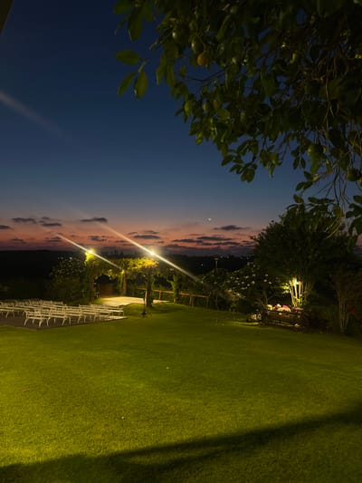This is a photograph taken in Beit Hanan, Israel, at dusk. The sky displays a gradient of deep blue at the top, transitioning to hues of orange and pink near the horizon, with scattered clouds. The foreground is dominated by a well-maintained, brightly lit green lawn. To the left, rows of white chairs are arranged on a wooden platform, suggesting an outdoor event setup.
In the middle ground, a raised platform with a white surface is visible, illuminated by overhead lights. The area around this platform is decorated with trellises and foliage. Further to the right, a tree is lit by a spotlight, highlighting its branches and a rustic wooden cart filled with flowers. The background shows a silhouetted landscape with distant lights. The overall scene is tranquil and suggests a venue prepared for an evening gathering.