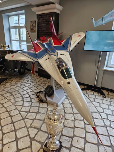A detailed view reveals a room's interior, seemingly a space within a museum, given the presence of a model fighter jet airplane of the MiG-29 Fulcrum variant, adorned with Soviet-era markings and prominently displayed along with a trophy. The model is affixed to a mount against a patterned, cobblestone-like textured wall.
The setting includes visible windows, a television screen, and additional, less distinct objects in the background, suggesting a curated exhibit. The overall lighting is bright, contributing to clear visibility of the artifacts. The location is assumed to be Архангельское, Russia, as per the prompt.