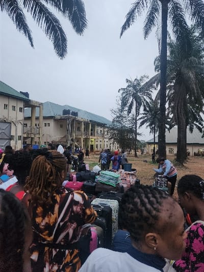 A crowd of people with luggage gathers in front of a multi-story building with a green roof and several water tanks on top. Palm trees flank the scene, reaching towards a cloudy sky. Many individuals appear to be women, some with distinctive hairstyles. The luggage consists primarily of suitcases and bags, suggesting travel or relocation. The overall atmosphere suggests a gathering or assembly, potentially at a transit point or lodging facility.