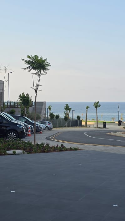 The image captures a parking lot scene in Agios Epiktitos, Northern Cyprus, with the sea visible in the background. In the foreground, a dark grey tiled area with embedded circular lights suggests a modern outdoor space. A curved asphalt road with double yellow lines marks a boundary. To the left, several parked vehicles, including black vans and a silver car, are visible. A few orange traffic cones are placed near the vehicles. A small garden bed with green plants and some pink flowers runs along the edge of the parking lot. A slender tree with sparse green foliage stands prominently in the mid-ground, with more trees and a set of stairs leading to a building in the background. The sea, a calm blue, stretches across the horizon, meeting a pale, hazy sky. Two figures, one in black and another in dark clothing, are walking on a pathway to the right, near a lamppost. The overall scene appears to be an urban setting near the coast, likely a public or commercial area.