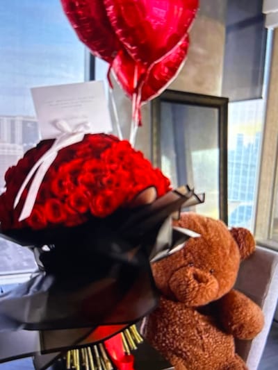 A close-up, blurred, indoor photograph captures a bouquet of red roses, heart-shaped red balloons, and a brown teddy bear arranged near a window. The window provides a distant view of a city skyline. The scene is dimly lit, with the focus on the floral arrangement and the teddy bear. There are no people visible in the image. The photograph does not provide any information about the city of Beșalma, Moldova.