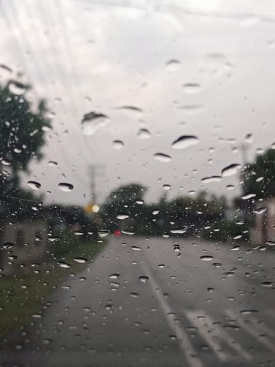 The image depicts a view through a rain-streaked car windshield. The scene outside is blurred due to the water droplets on the glass. A road is visible, with trees and vegetation lining its sides. There are some streetlights and traffic signals in the distance, suggesting an urban or semi-urban environment. The sky is overcast and gray, indicating a rainy day. The location is identified as Bale Musara, Indonesia. No people or specific actions are discernible.