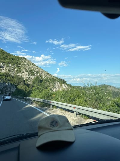 The image depicts a view from inside a vehicle traveling on a road adjacent to a mountainside. The sky is blue with scattered clouds. The road is paved and has a guardrail. Vegetation covers the mountainside. A baseball cap is visible partially obscuring the view through the vehicle's window. A vehicle is visible in the distance on the road. Location is likely in or near Dobrsko Selo, Montenegro based on the landscape.