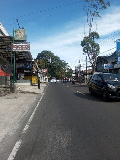 The image depicts an outdoor street scene under a clear blue sky with scattered white clouds, situated in the city of Phonengam, Laos. The perspective is from a low angle on the left side of a paved asphalt road, looking forward down the street.

On the left, a concrete sidewalk with a visible drain grate leads to a commercial building featuring a black metal gate and a cantilevered roof. Attached to this structure are multiple signs, including a green sign partially showing "SBN," and a brown sign stating "PRAKARSA JAYA STEEL" and "SUPPLIER BESY & BAJA" (Supplier of Iron & Steel), along with phone numbers (022-86861871 and 081223157811). One person, dressed in a dark uniform, stands on the left sidewalk near the road's edge, facing left. Additional buildings and foliage are visible further down the left side.

The asphalt road, dark gray and showing minor wear, occupies the center of the frame. A solid white line delineates the left edge of the road, and yellow dashed lines are visible in the distance, marking traffic lanes. Traffic includes a black multi-purpose vehicle (MPV) partially visible on the right side in the foreground, facing away from the viewer. Further down the road, multiple vehicles, including white cars and numerous motorcycles and scooters, are present, with a cluster of motorcycles approaching.

On the right side of the road, a tall, slender tree with sparse foliage stands. Several utility poles with overhead wires line this side of the street. Beyond these, commercial buildings with signage, such as one displaying "73 CUC HILFING," extend into the background. Dense green trees are visible in the far background. Shadows are cast across the road and structures, indicating direct sunlight.