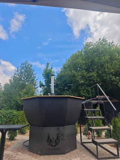 A wide shot captures an outdoor scene with a large, black, octagonal hot tub as the central subject. The hot tub is made of metal and has a wooden rim. It sits on a base with the word "GORISTA" and a flame-like logo etched into it. A silver chimney extends from the top of the hot tub. To the right of the hot tub, a metal staircase with wooden steps leads up towards the rim. In the background, a variety of green trees and bushes are visible, along with a blue sky with scattered white clouds. A portion of a table is visible in the lower left corner of the image, and a brick-like paved area forms the ground. The overall setting appears to be a backyard or garden area, suggesting it is a private residence in the city of Khlevino, Russia. There are no people visible in the image.
