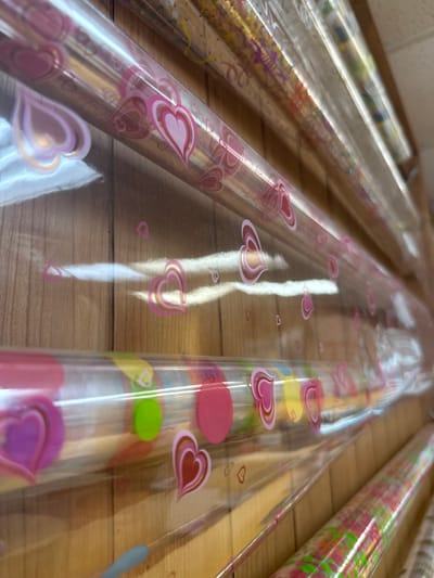 The image shows a close-up view of several rolls of wrapping paper with different patterns. The rolls are arranged horizontally on what appears to be a wooden shelf or surface. The focal point is on the middle roll, which features a clear plastic wrapping with pink heart designs. Other rolls with varied colors and patterns are partially visible on either side. The scene is indoors, likely in a store or storage area. The location is Davie, United States of America.