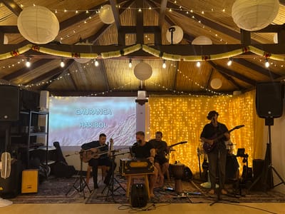 A band performs in an indoor space with a thatched bamboo ceiling, decorated with paper lanterns and string lights. The stage is set up with musical instruments including guitars, a harmonium, a djembe drum, and speakers. Four men are visible, three of whom are playing instruments. The man on the far left plays an acoustic guitar, the man in the center plays the harmonium and sings into a microphone, and the man on the far right plays an electric guitar. The fourth man is partially obscured behind the electric guitarist. A projector screen behind the band displays text that reads "GAURANGA HARIBOL" in a light blue gradient. The overall atmosphere appears to be a performance or concert. The time of day is not clearly indicated, but the indoor lighting and string lights suggest an evening event. The location is Tong Yang, Thailand, as indicated by the context.