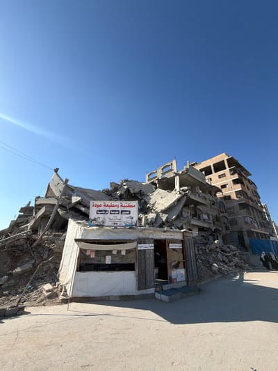 In Jabalia, Palestinian Territories, the aftermath of destruction is evident. A partially collapsed multi-story building dominates the scene, with concrete debris piled high. In front of the damaged structure, a small, temporary-looking shop with a white tarp roof stands, displaying signs in Arabic that read "Maktaba wa Matba'a Ouda" (Ouda Bookstore and Printing). The shop appears to be functional, with goods visible inside and a wooden door ajar. To the right of the damaged building, another, more intact multi-story structure with visible windows stands. Two figures, dressed in dark clothing and headscarves, are seen walking in the background. The ground is a dusty, paved surface, and the sky above is clear and blue. Power lines are visible strung across the sky.