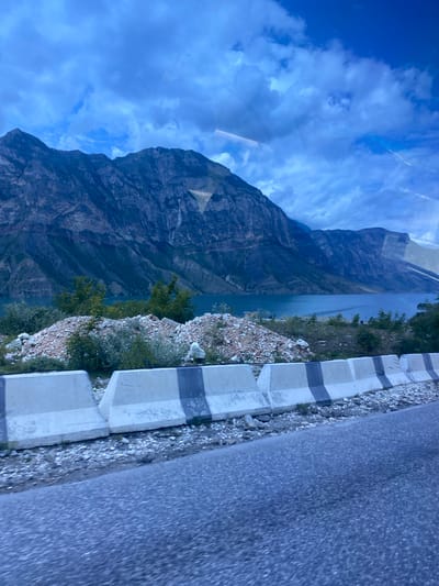 The image depicts a scenic mountain landscape with a body of water, likely the Irganayskoye Reservoir, in the foreground. A road runs along the edge of the water, separated by concrete barriers with black stripes. Mountains rise steeply behind the water, their slopes covered in vegetation and rugged terrain. The sky is cloudy, with hints of blue peeking through. The overall impression is one of natural beauty and a remote, mountainous region. The presence of the road and barriers suggests human infrastructure has been integrated into this natural environment.