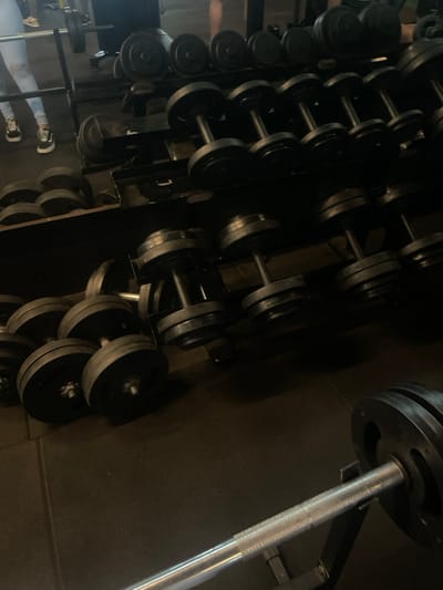 The image displays the interior of a gym located in Anápolis, Brazil, featuring dark, possibly rubberized, flooring. The scene is dominated by various weightlifting equipment. Multiple sets of black dumbbells with metallic handles are systematically organized on multi-tiered, dark metal storage racks that span the mid-ground and background. Additional dumbbells are observed on the floor in the left foreground and mid-ground. A silver barbell with black weight plates is prominently positioned horizontally on a black stand in the immediate foreground. To the far left in the background, a person's lower body is visible, clad in light-colored athletic leggings and dark sneakers with white soles, standing near a weight bench and another barbell. A faint, indistinct reflection of another individual's lower extremities is discernable in the upper background, likely in a mirror. Artificial lighting illuminates the area, producing some shadows.