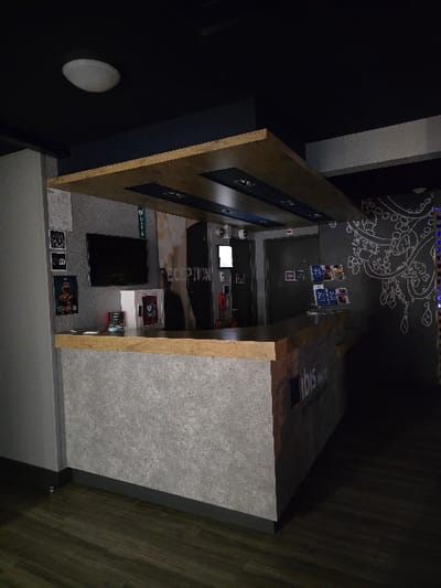 The scene shows a high-angle, slightly rotated view of an interior. A cabinet with a wooden frame and a light grey exterior is positioned against a dark wall. Inside, a counter with embedded screens and various objects is visible. The top of the cabinet is branded "ibis". The backdrop features a grey wall with white, abstract drawings. A white, round object is on the dark wall.
