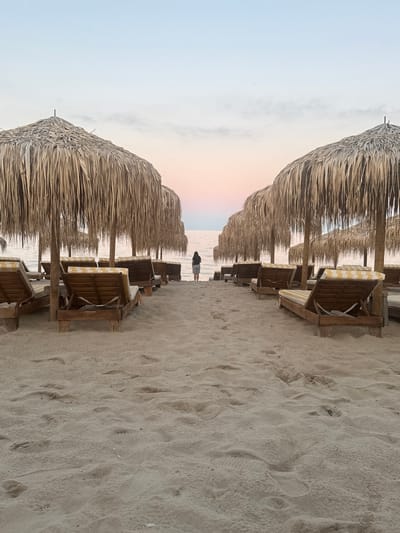 In Kranevo, Bulgaria, on a sandy beach at dusk, two rows of empty, sun-bleached thatched umbrellas face the sea. Beneath the umbrellas are wooden lounge chairs with yellow and white striped cushions. A lone figure, seen from the back, stands at the water's edge between the rows of umbrellas, facing the calm sea and the pastel-colored sky. The sand in the foreground is imprinted with footprints.