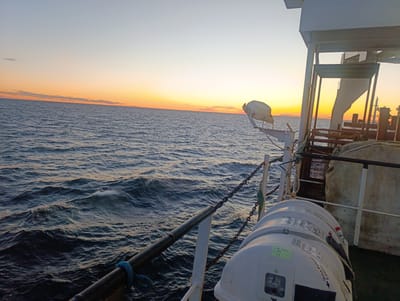 The image shows a view from the deck of a vessel at sea, with a sunset creating a warm orange glow on the horizon. The choppy, dark blue water fills the majority of the frame. In the foreground, a white cylindrical object, identified by a green sticker as "Sea Project - 3661 2/2," is mounted on the deck. A metal railing and a thick chain run along the edge of the vessel. On the right side of the image, part of the vessel's structure is visible, including a white railing, some metal supports, and what appears to be a large, light-colored cover. The sky is a pale blue, transitioning to orange and yellow near the horizon. The scene is devoid of people and any discernible city landmarks. The location is the sea, not within the city of Larsmo, Finland.