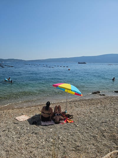 The image depicts a sunny day at a pebble beach in Jošice, Montenegro. In the foreground, two individuals are seated on towels under a colorful beach umbrella. One person is facing away from the camera, wearing a black bikini, while the other is lying down, facing towards the water.

The water is clear and calm, with small waves lapping at the shore. In the distance, there are several rows of oyster farms or similar aquaculture structures. A few people are visible in the water, one person on an inflatable raft and another swimming. A passenger boat is sailing in the background. The sky is a clear, vibrant blue. The overall atmosphere is one of relaxation and enjoyment of a summer day at the seaside.