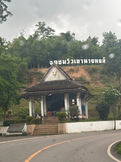 The image shows a road leading to a small, open-sided building situated on a hillside covered in greenery. A white sign with Thai lettering is prominently displayed on the slope above the building. The building has a dark tiled roof with a triangular facade featuring a golden bird emblem. Steps lead up to the building from the road. The sky is overcast. The location is Ban Pak Ra, Thailand.