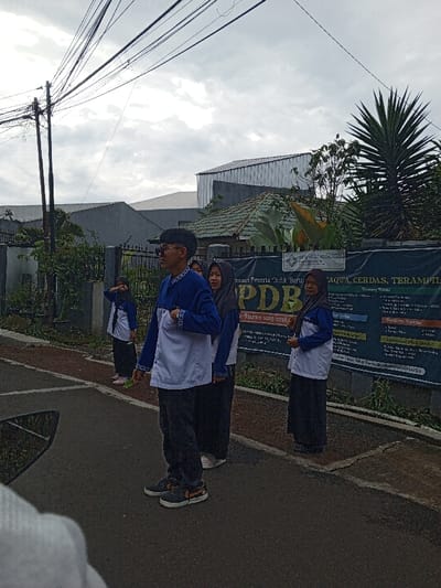 A group of four young people wearing matching blue and white outfits stand in a row on the side of a street. The first person is a boy, and the other three are girls with hijabs. Behind them is a fence with a banner that reads "SKPLAADOA SAH imaan Peserta Didik Baru PDB in Haran 2025-2025 AQWA, CERDAS, TERAMPIL." The banner also includes text about school activities. The sky is cloudy, and there are some buildings in the background.