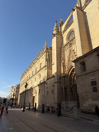 This media file depicts a low-angle, sunlit view of a grand, ornate cathedral facade and its surrounding street in Seville, Spain. The architecture is a blend of Gothic and Renaissance styles, characterized by intricate stonework, pointed arches, and decorative carvings. Sunlight casts long shadows, indicating it's likely late afternoon or early evening.
Several people are visible walking along the cobblestone street in front of the cathedral. Some are strolling casually, while others appear to be engaged in conversation. The street is lined with lampposts and chains, suggesting a public thoroughfare. In the distance, other buildings can be seen, contributing to the urban setting.
The sky is a clear, vibrant blue, indicating fair weather. The overall atmosphere is one of a peaceful, lively urban environment, with the magnificent cathedral serving as a prominent backdrop. The image conveys a sense of awe and grandeur due to the scale and detail of the architecture. There is no visible text in the image.