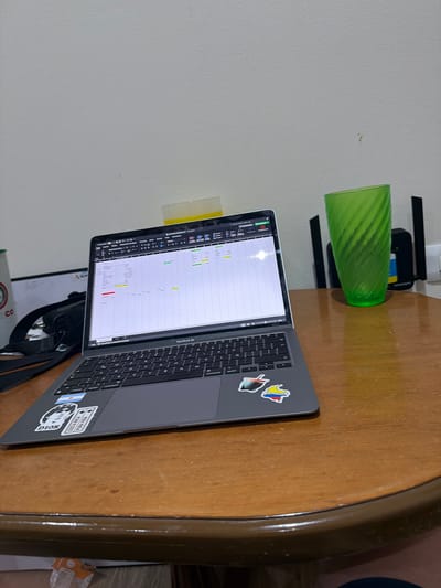 The image depicts a close-up of a wooden table with a MacBook Air laptop placed on it, open and displaying a spreadsheet. To the right of the laptop is a green, ribbed plastic cup, and further to the right, partially visible, are two black objects that appear to be part of a wireless router. In the background, a plain, light-colored wall is visible. The laptop has several stickers on its surface, including one with the flag of Argentina, another with a stylized image of Diego Maradona, and one depicting the map of Colombia. The lighting suggests an indoor environment. The scene is situated on a table that appears to be in a domestic or office setting. There is no explicit visual information within the image to determine the location as Cancún, Mexico.