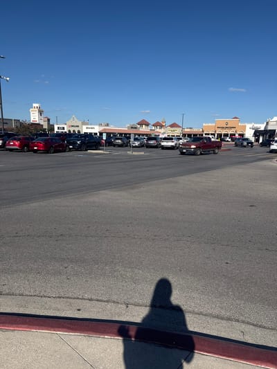 A street scene on a sunny day, featuring parked cars lining buildings with various retail signs. The sky is clear and blue. A wide asphalt road stretches into the distance, marked with faint lines. A red curb separates the road from a sidewalk where a person's shadow is cast.