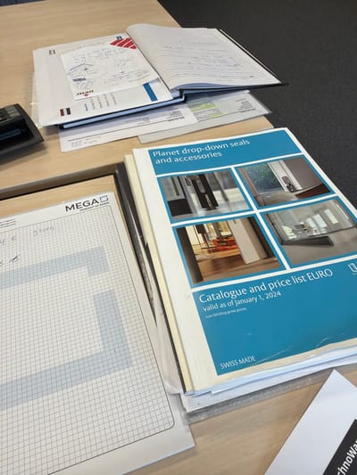 The image displays a static, overhead view of a section of a light-colored wooden desk in an office or workspace environment. A dark gray carpet is visible in the background.

On the left side of the desk, the partial top-left corner of a black electronic device, possibly a calculator, is visible. Adjacent to it, a stack of documents includes a white sheet with a red and blue "DENI" logo and handwritten notes on a smaller white paper overlay. Beneath this is an open, black-bound ruled notebook with handwritten text.

In the bottom-left quadrant, a white sheet of grid paper with the "MEGA PERFECT IN FORM" logo is present, featuring handwritten notes that include "Drog" and euro symbols alongside numbers.

Towards the right side, a prominent stack of catalogues is observed. The top catalogue features a blue cover with white text reading "Planet drop-down seals and accessories." Below this title, four image squares illustrate various door seal applications. Further down the cover, text states: "Catalogue and price list EURO valid as of January 1, 2024 non-binding gross prices," with "SWISS MADE" at the bottom.

A small corner of a white paper with partially visible black text, potentially "TechnoWar," is visible in the bottom-right corner.

No individuals or actions are depicted in the scene. The image provides no visual information to identify a specific location within the city of Apeldoorn, Netherlands.