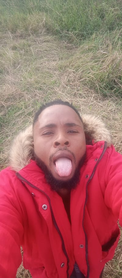 A selfie shows a man lying in dry grass wearing a red hooded winter coat with fur trim. He faces the camera with his tongue sticking out. He has a beard and short hair. The background is a field of dry grass and some green grass. The photo is taken from a high angle.