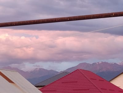 The image captures a view from a residential area, likely in Іргелі ауылдық округі, Kazakhstan, during a twilight hour. In the foreground, several rooftops are visible, including one with a bright red, textured surface, another with a dark gray, textured surface, and a third partially visible white roof with an orange accent. A thick, rusty metal pipe runs horizontally across the upper portion of the frame, casting a shadow. A thin, white wire also stretches diagonally across the sky. In the background, a mountain range with snow-capped peaks is silhouetted against a dramatic sky filled with clouds tinted in shades of pink, purple, and blue, indicative of a sunset or sunrise. The overall scene suggests a peaceful, rural or semi-urban landscape with a backdrop of natural grandeur.
