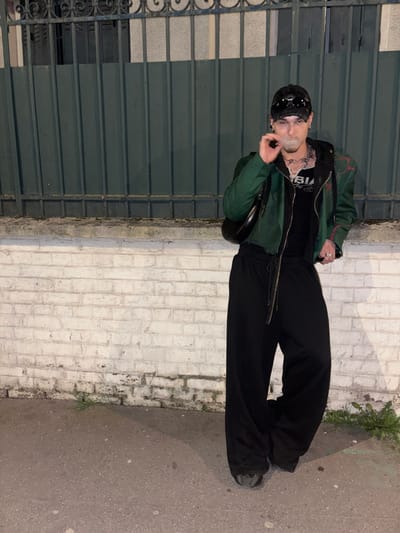 A full-body photograph depicts a single individual standing on a paved surface in Asnières-sur-Seine, France. The individual, appearing male, is positioned centrally, facing the camera directly. They wear a black baseball cap, dark sunglasses, a dark green jacket, a black hooded garment, and a black tank top with white lettering. Multiple silver-colored chains are visible around the neck, and rings adorn the fingers of the right hand. The individual wears baggy black trousers with a visible drawstring and dark, possibly chunky, shoes. A black crescent-shaped bag is slung over their left shoulder. The individual holds a lit cigarette or similar object between the thumb and forefinger of their right hand, with a plume of smoke emanating from their mouth. Behind the individual is a low white brick wall, exhibiting some discoloration and minor vegetation growth at its base. Above this wall is a dark green metal fence or gate composed of vertical bars and a decorative top rail. The ground is dark asphalt or concrete, with minimal plant growth visible. The scene is illuminated by artificial light, suggesting a nighttime setting.