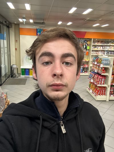 A young man in a black hooded jacket poses for a selfie indoors, likely in a store. A rack filled with packaged snacks is visible behind him. The lighting is bright and fluorescent. His hair is light brown, and he has a light beard. He looks directly at the camera with a neutral expression. The scene appears to be a convenience store or a similar retail environment.