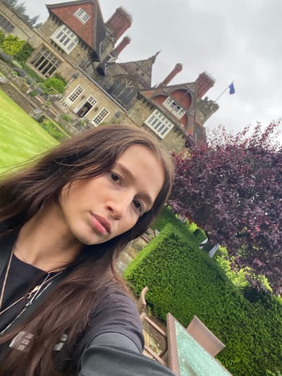 In Chichester, United Kingdom, a young woman with long brown hair is pictured in a selfie. She is wearing a black t-shirt and several necklaces. Behind her, a large, ornate building with red-tiled roofs and a blue flag on a pole is visible. The building appears to be a historic manor or castle, surrounded by a meticulously manicured garden with neatly trimmed hedges and a vibrant red-leafed tree. The sky is overcast, suggesting a cloudy day.