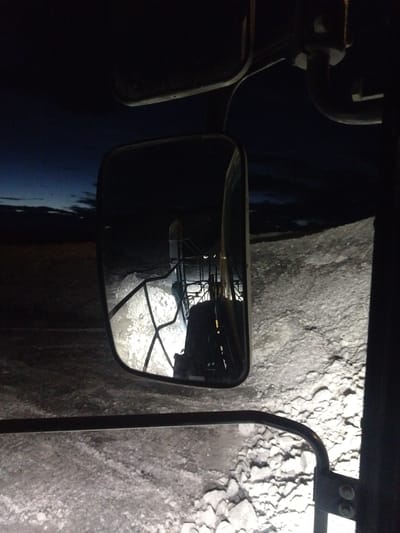 A rearview mirror reflection shows a person in a dark environment, likely operating heavy machinery. The reflection reveals a metal ladder and a control panel with what appears to be a backpack hanging from it. The ground visible in the reflection is covered in light-colored debris, possibly snow or gravel. The original image shows a dark, possibly nighttime scene, with the headlights of the vehicle illuminating the ground in front. The setting appears to be outdoors, possibly in an industrial area or a construction site in Naminga, Russia, given the limited visibility and the presence of what looks like construction materials. The overall atmosphere is stark and industrial.