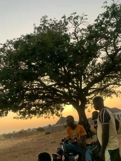 This outdoor photograph, taken in Burji, Nigeria, captures a scene at either sunrise or sunset, indicated by the warm, golden light bathing the sky and landscape. A large, mature tree with dense green foliage dominates the left side of the frame, its branches reaching upwards.

On the right side of the image, several individuals are gathered on bare, dusty ground. In the midground, a man wearing a yellow and black striped t-shirt stands, facing towards the left. Beside him, another man is seated on a motorcycle; he wears an orange t-shirt featuring a graphic of a cartoon bear. Other figures are partially visible behind these individuals, some obscured by the tree or the warm lighting.

The background reveals a hazy, golden horizon with the faint outlines of simple buildings or structures in the distance. The ground appears dry and unpaved, consistent with a rural or semi-urban environment. The overall atmosphere is tranquil, with people gathered casually under the tree during the soft light of dawn or dusk. No distinct readable text is visible other than the graphic on the orange t-shirt.