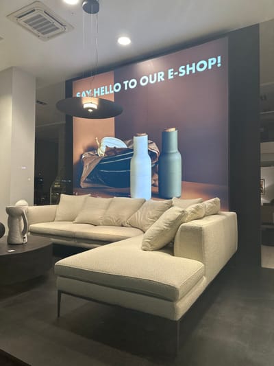 The image depicts an indoor commercial setting, likely a modern furniture or lifestyle showroom, located in Engomi, Nicosia, Cyprus. The primary subject is a contemporary, light beige or off-white sectional sofa with multiple matching throw pillows, suggesting comfort and style.

Behind the sofa, a large vertical display screen serves as a prominent advertising feature. The screen showcases two insulated bottles with wooden caps: one in a dark teal or green color, and the other in a light blue or white shade, displayed against a dark, textured background. The screen also features white, capitalized text that reads: "SAY HELLO TO OUR E-SHOP!".

In front of the sofa, a dark, round coffee table holds a small, light-colored decorative object, possibly a jug or vase. Above, a sleek black pendant light hangs from the ceiling, contributing to the modern aesthetic. An air conditioning unit is visible on the ceiling to the left. The space appears well-lit by artificial lights, making the time of day undeterminable. No individuals are present in the scene, which is arranged to highlight the displayed products and create an inviting atmosphere.