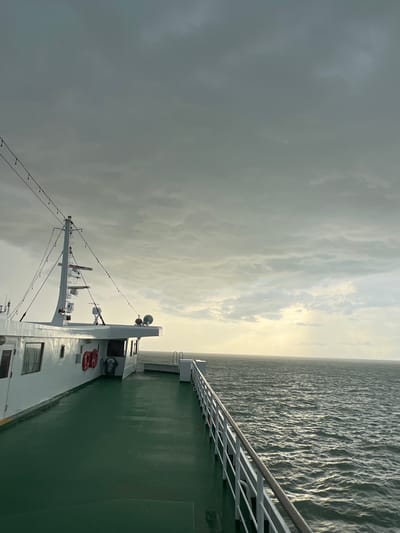 An exterior, wide shot captures a view from the deck of a ferry in Don Sak, Thailand. The scene is overcast with dramatic, gray clouds filling the sky, but a sliver of sunlight breaks through on the horizon, illuminating the choppy, dark sea. The ferry's white superstructure is visible on the left, with a green deck leading to a white railing. Life preservers are mounted on the side of the ferry. The water is textured with numerous small waves, and the overall mood is somber, possibly indicating approaching bad weather.