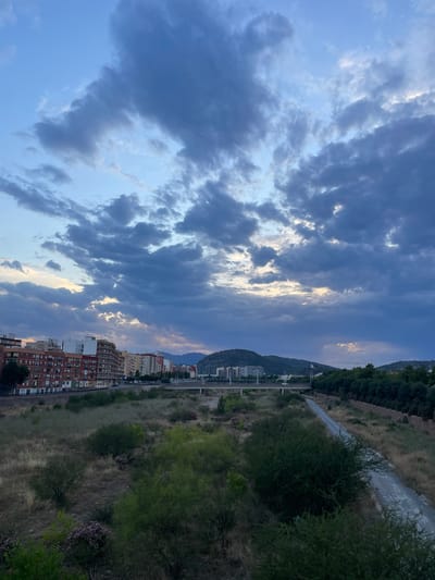 The image depicts a view of Sagunto, Spain, at dusk. A dense cluster of apartment buildings lines the left side of the frame, with a road running in front of them. Beyond the buildings, a bridge spans across a dry riverbed or floodplain, where dry grass and scattered bushes dominate the landscape. To the right, a gravel path winds its way through overgrown vegetation. In the distance, rolling hills covered in trees form the backdrop, with a few more buildings visible on their slopes. The sky is filled with dramatic, dark clouds, tinged with the warm hues of sunset peeking through gaps. The overall scene suggests an urban fringe environment experiencing the transition from day to night.