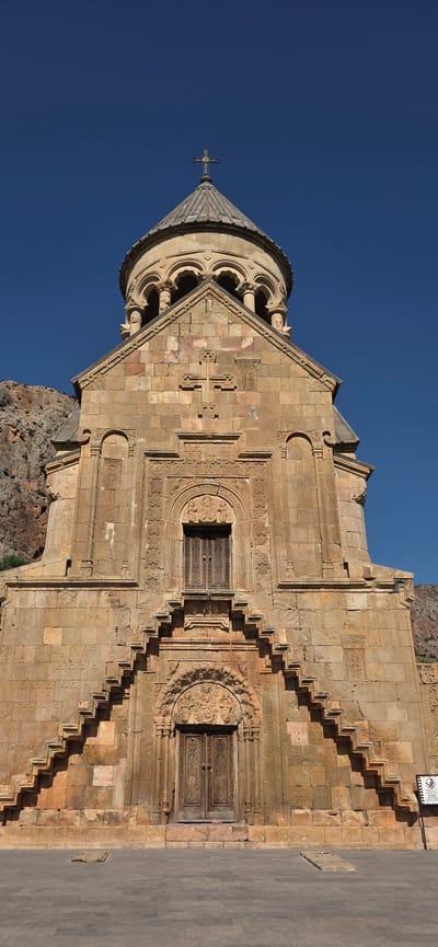 The image shows a low-angle, full frontal view of the Noravank Monastery church facade, located in the city of Amaghu, Armenia. The church is constructed of ochre-colored stone and features a prominent, conical dome topped with a cross. The facade is intricately carved with religious motifs, including a large cross above a decorative arched window with wooden doors. Below this window, a steep, zigzagging stone staircase leads to another set of ornate wooden doors, above which is a carved tympanum depicting figures. The church is situated against a backdrop of rugged, rocky cliffs under a clear blue sky. To the right of the entrance, a small, square sign with symbols is visible. The foreground consists of a paved courtyard. No people are present in the image.