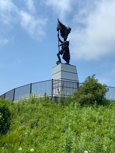 A statue of a soldier holding a flag aloft stands on a white pedestal, which is surrounded by a black metal fence. The statue and pedestal are situated on a grassy hill in Kraskino, Russia. The sky is blue with scattered white clouds. The hill is covered in green grass and some wildflowers.