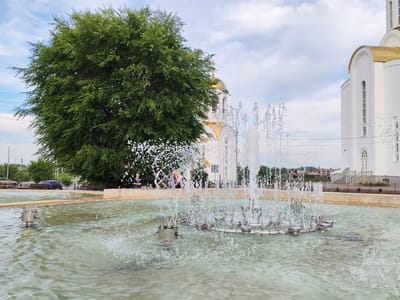 This image captures a public fountain with multiple water jets actively spraying water. In the background, a large, dense green tree dominates the left side of the frame, partially obscuring a white building with a golden dome and cross, characteristic of a Russian Orthodox church. The building has arched windows and decorative elements. The fountain is situated in a shallow pool of water with a light green tint. Several individuals are visible standing or sitting near the edge of the fountain area, some partially obscured by the water spray. In the distance, a street with cars and other buildings can be seen. The sky is partly cloudy. This scene is located at the fountain complex in the city of Kurgan, Russia, specifically at the central square near the Alexander Nevsky Cathedral.