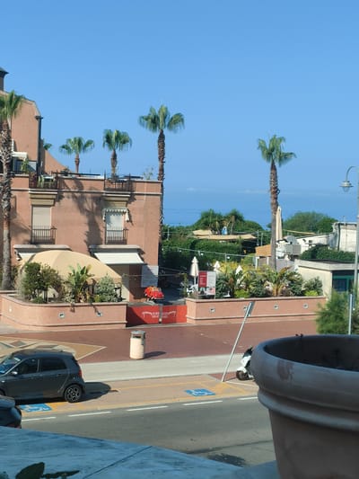 The image captures a street-level view in Genoa, Italy, under a clear blue sky. In the foreground, a large, textured terracotta planter dominates the right side of the frame. To the left, a road is visible with parked cars, including a dark gray hatchback with its rear facing the viewer. A scooter with a white body and dark accents is parked on the side of the road.
Further into the scene, a building with a reddish-brown facade features multiple balconies with black railings. Several palm trees are planted around and on top of the building, adding to the Mediterranean ambiance. A large beige umbrella shades an outdoor seating area at the base of the building, where lush green plants are also present. A small, bright red toy car is placed on a red platform in front of the building. Beyond this, more greenery and smaller structures are visible, suggesting a commercial or hospitality area. In the distance, the shimmering blue expanse of the sea can be seen under the bright sky. A street lamp stands tall on the right side of the image.