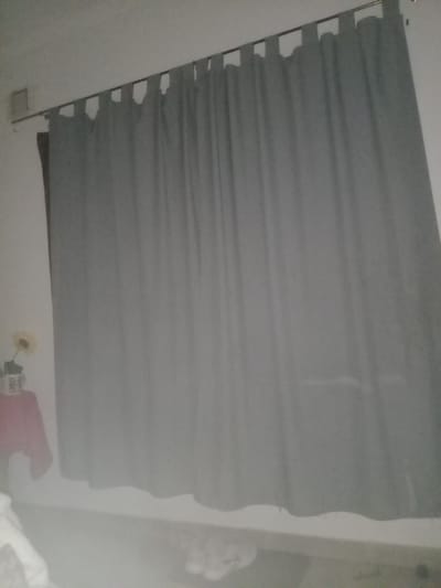 The image shows a gray fabric window shade hanging in a room. The shade has a series of evenly spaced slits along its left side, likely for a pull cord or attachment mechanism. The wall is white, and there is a hint of a room's interior below the bottom edge of the shade. Faintly visible are hints of a floral decoration, a pink item, and some white soft materials (possibly blankets or clothing), all of which sit on a flat surface. Details are insufficient to locate the exact address within Villa Ballester Noroeste, Argentina. No people are visible. The environment is indoors, suggesting residential or domestic use. The objects include a window shade, wall, and hints of domestic items. The actions are implicit; the shade is hanging and possibly designed to control light or privacy. Interactions are not directly observable.