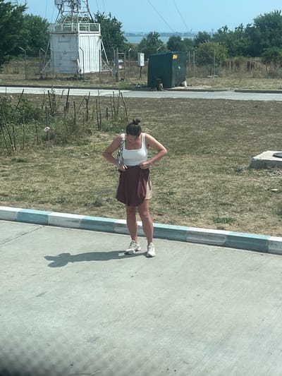 A woman stands on a concrete surface in Ovidiu, Romania, adjusting her maroon and tan skirt. She is wearing a white tank top and white sneakers. Her hair is pulled back in a bun. Behind her, a dry, grassy field is bordered by a curb with blue and white accents. In the background, there is a large white structure with antennas, a green portable building, and trees. The sky is clear and blue.