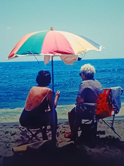 Two individuals are seated on chairs on a beach under a colorful beach umbrella. The person on the left is facing away from the camera, wearing a black swimsuit. The person on the right, also facing away, is wearing a patterned shirt and sunglasses. In front of the person on the right, a red life vest is attached to their chair. The ocean is visible behind them, with the sky above. The location of this scene is not Vitoria-Gasteiz, Spain, as the image depicts a beach, and Vitoria-Gasteiz is an inland city.