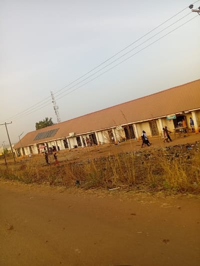 The image captures an outdoor scene, likely a street or unpaved road in a semi-urban or rural setting.

In the foreground, a wide, reddish-brown dirt road runs diagonally from the lower left to the lower right corner. The road appears unpaved and has some faint marks or indentations on its surface.

Separating the road from the buildings in the midground is a strip of dry, tall, yellowish-brown grass and sparse weeds. Scattered within this grassy area are pieces of litter and debris.

The midground is dominated by a long, low-rise building with a light-colored (possibly off-white or cream) facade and a continuous, sloping reddish-brown or terracotta-colored roof. The roof appears to be made of corrugated metal or tiles. The building is divided into multiple units, each with a doorway, some of which have open fronts or small stalls set up in front of them. Towards the left side of the building, several dark rectangular panels, possibly solar panels, are visible on the roof.

Several people are scattered along the front of this building. On the left side, two or three individuals are visible, one holding a red umbrella open, possibly for shade from the sun or as a means of transport. Further to the right, more individuals are standing, walking, or seated near the shop fronts, suggesting activity like commerce or social interaction. Some small tables or display stands are visible in front of a few units. One unit, towards the right, has a small green canopy or awning extended over its entrance.

In the background, a pale, hazy, light gray sky covers the upper half of the image. Power lines stretch diagonally across the sky from left to right. On the far left, a utility pole with power lines is visible, and further back, a tall metal lattice transmission tower or communication mast stands against the sky. A few distant trees or bushes are barely visible on the horizon to the left.

The overall impression is of a bustling but modest commercial or residential area, with the soft, diffused light suggesting either early morning or late afternoon, or an overcast day. No legible text is visible in the image.