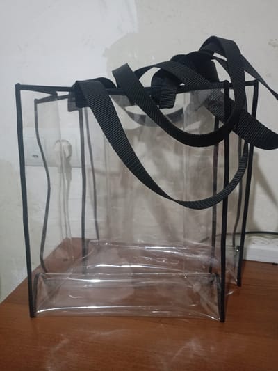 The scene is located in the city of Koghb, Armenia. Two transparent, rectangular tote bags, constructed from clear plastic with black fabric handles and matching black trim along all edges, are positioned upright on a reddish-brown wooden surface. One bag is slightly in front of and to the left of the other. The black fabric handles are gathered at the top. In the background, visible through the transparent bags, is a light-colored, textured wall. A white electrical outlet with a white power plug inserted into its top socket is visible on the wall, with a white cable extending downwards. To the right of the outlet, a white multi-socket power strip is partially visible. The wooden surface exhibits reflections. No individuals are present in the image.